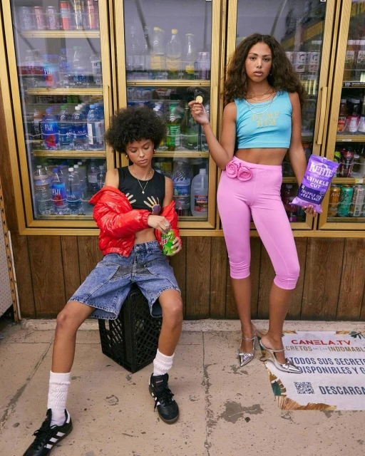 Two stylish women pose indoors near glass refrigerators. One sits on a crate, holding a soda, while the other holds chips. 