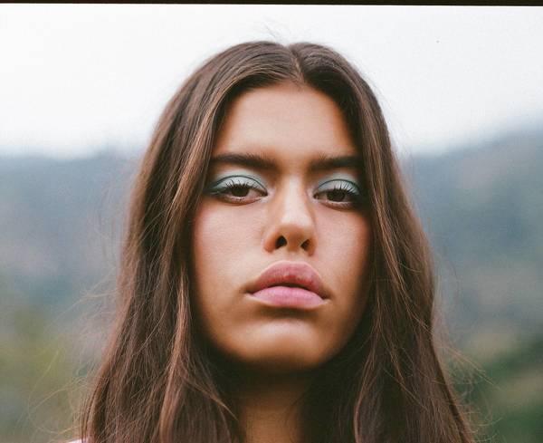 Close-up portrait of a young woman with long brown hair and teal eyeshadow, set against an out-of-focus green and grey landscape.