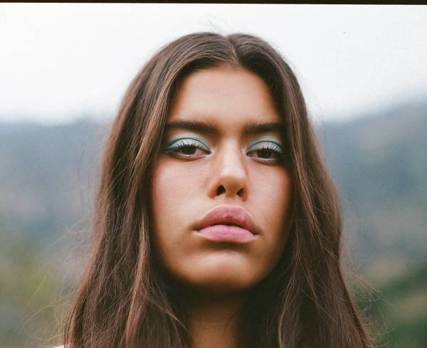 Close-up portrait of a young woman with long brown hair and teal eyeshadow, set against an out-of-focus green and grey landscape.