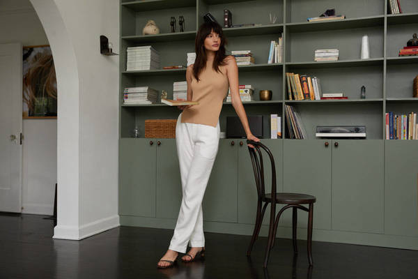 A woman in a beige top and white pants stands in a room with a sage green bookshelf and dark hardwood floors, holding a book.