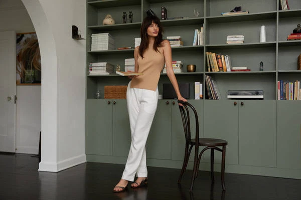 A woman in a beige top and white pants stands in a room with a sage green bookshelf and dark hardwood floors, holding a book.