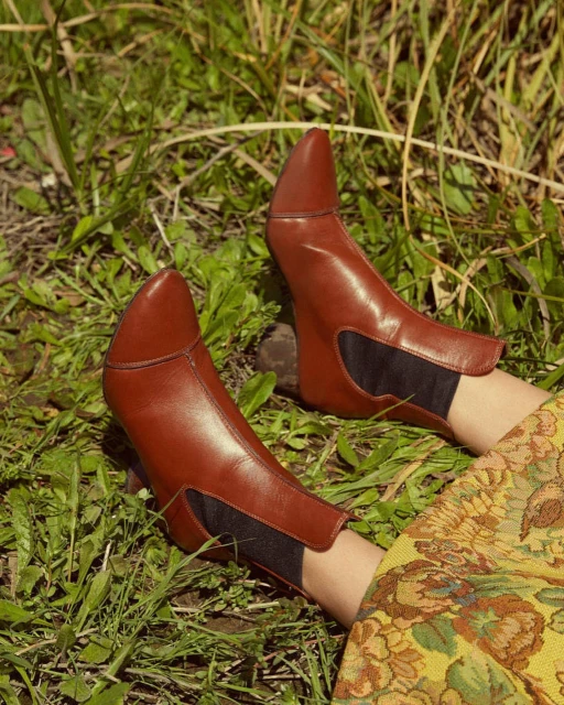 A pair of tan, pointed-toe leather heeled ankle boots are set on grass near a yellow floral-patterned dress in an outdoor setting.