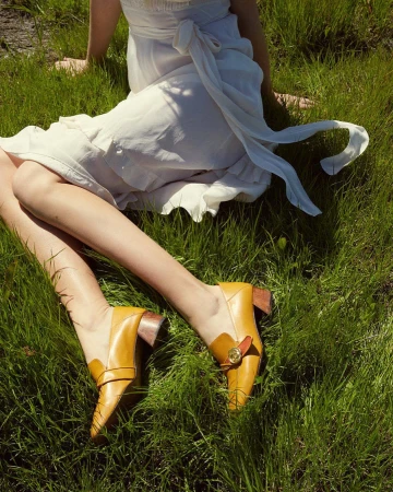 Close-up of a seated woman wearing a white dress and yellow leather loafers resting on vibrant green grass.
