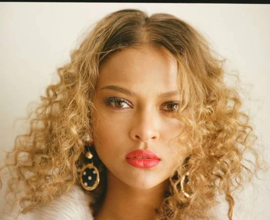 Close-up portrait of a woman with voluminous curly blonde hair, red lipstick, and large circular earrings against a plain background