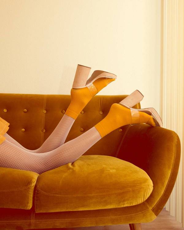 Person in beige fishnet tights and yellow socks lying on a mustard-yellow tufted couch with tan platform heels and a cream wall.