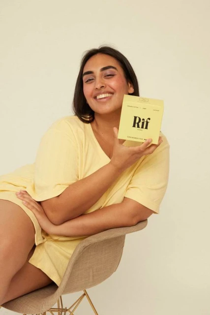 Smiling woman in yellow loungewear sits on a chair and holds a yellow package in a light-filled indoor setting, showcasing a relaxed and positive vibe.