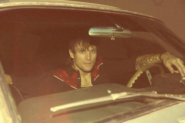 Adult male with a serious expression sitting inside a vintage car with a steering wheel in an indoor environment with warm, vintage-toned lighting.