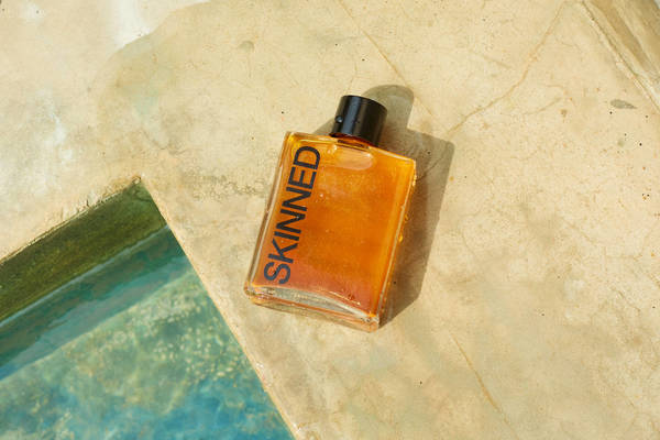 Glass bottle of orange liquid with black cap and 'SKINNED' text on a tan textured surface with a glimpse of turquoise water.