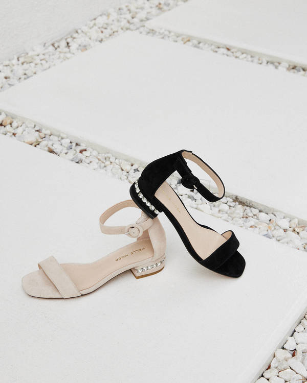 Two single-strap heeled sandals, one black and one beige, lay on a light gray and white tile surface with small white pebbles.