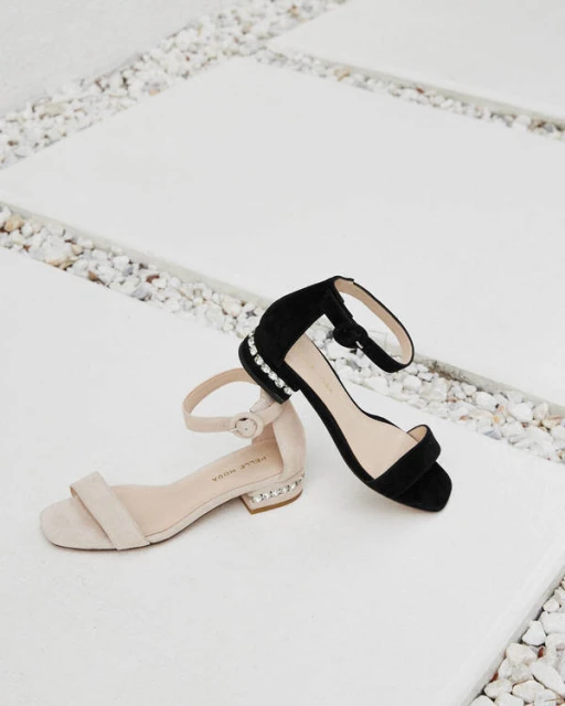 Two single-strap heeled sandals, one black and one beige, lay on a light gray and white tile surface with small white pebbles.