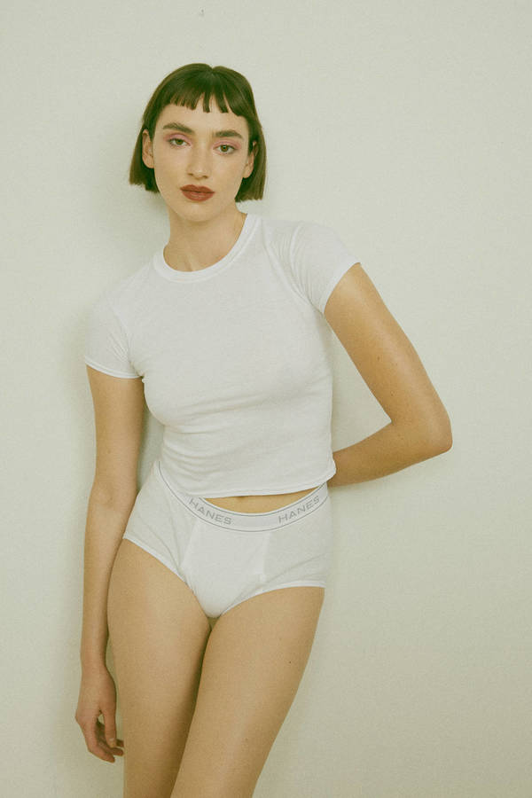 Indoor shot of a fair-skinned young adult woman with short brunette hair, wearing a white t-shirt and underwear, posing against a white wall.