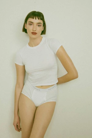 Indoor shot of a fair-skinned young adult woman with short brunette hair, wearing a white t-shirt and underwear, posing against a white wall.