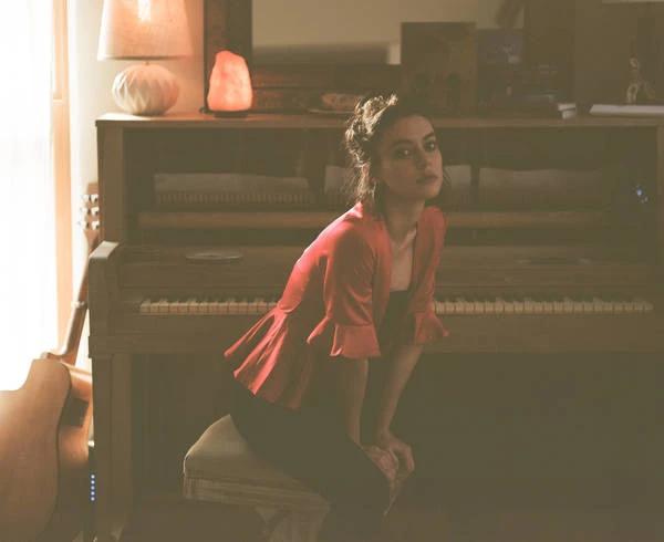 Young woman poses on a chair in front of an upright piano in a warmly lit room, wearing a red blouse and dark pants.