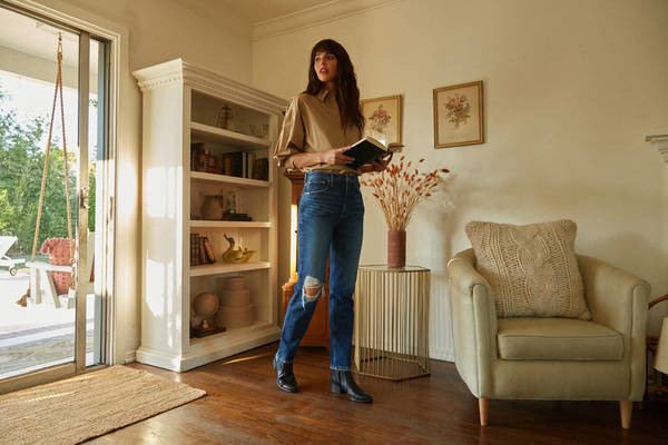 A woman with long brown hair stands in a living room, wearing jeans, a tan shirt, and black boots, holding a book near a bookshelf.