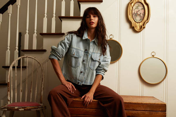A brunette woman with bangs in a denim jacket and brown pants sits on a wooden chest indoors with mirrors and stairs in the background.