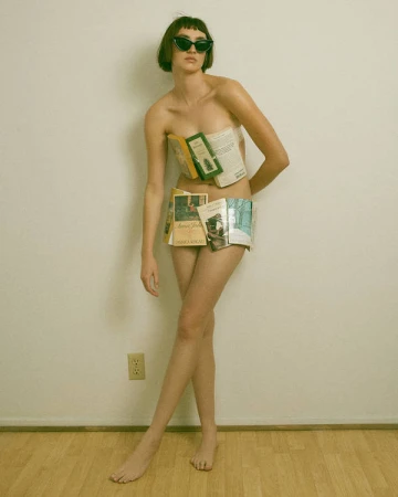Woman wearing cat-eye sunglasses and open books covering her torso, standing against a plain wall.
