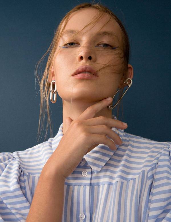 Close-up of a fair-skinned woman wearing a striped shirt with blue eyeliner, a large safety pin earring, and a silver metal earring.