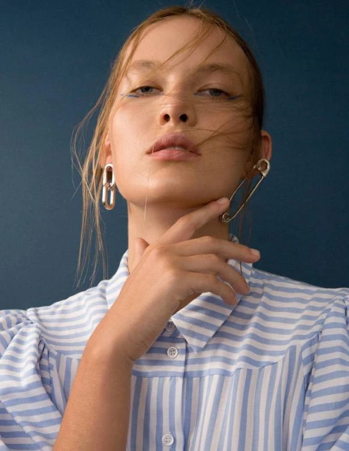 Close-up of a fair-skinned woman wearing a striped shirt with blue eyeliner, a large safety pin earring, and a silver metal earring.