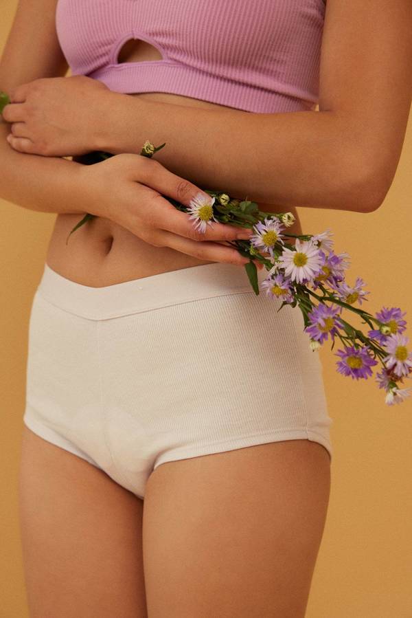 Mid-shot of an adult woman holding purple flowers in her hands, wearing a lavender top and white knit shorts against yellow background.