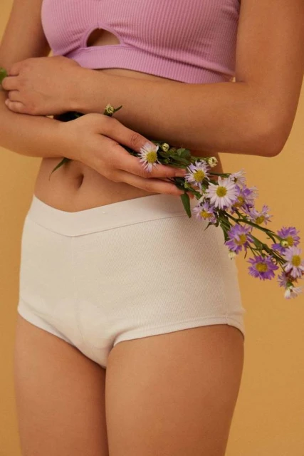 Mid-shot of an adult woman holding purple flowers in her hands, wearing a lavender top and white knit shorts against yellow background.