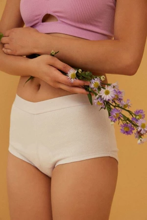 Mid-shot of an adult woman holding purple flowers in her hands, wearing a lavender top and white knit shorts against yellow background.