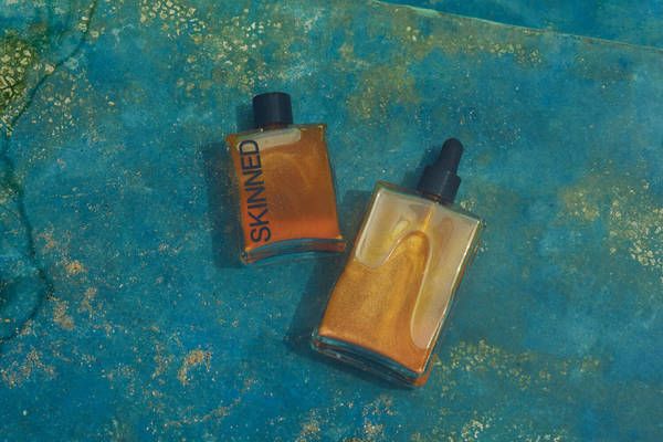 Two bottles of body oil with shimmer sit on a turquoise surface, one labeled 'SKINNED,' shot with overhead studio lighting. 