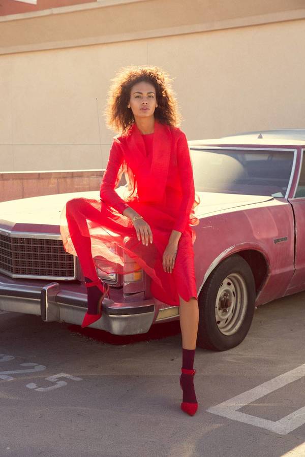 A young woman in a red dress poses against a faded pink vintage car in an outdoor lot with a neutral-toned building in the background.