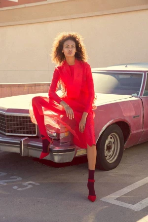 A young woman in a red dress poses against a faded pink vintage car in an outdoor lot with a neutral-toned building in the background.