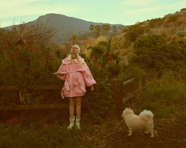 Woman in a pink quilted coat with a dog on a grassy path, with a mountain range and light blue sky in the background.