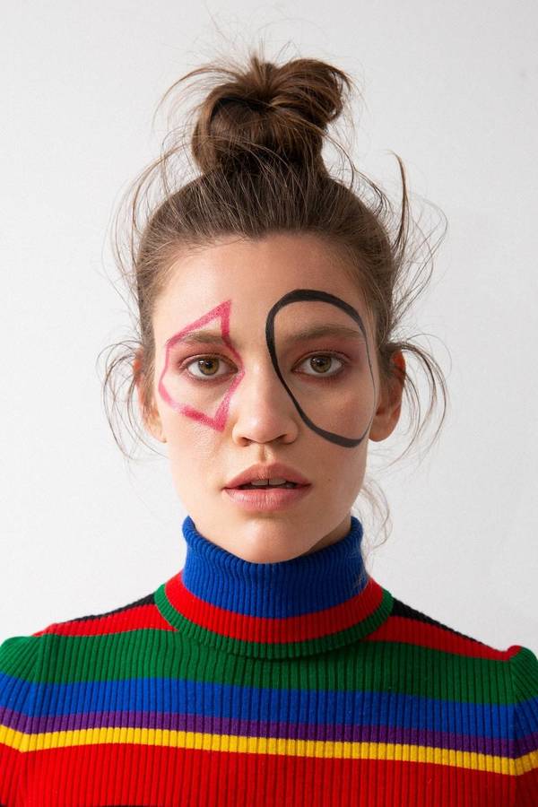Close-up of a woman with geometric makeup wearing a colorful striped turtleneck sweater against a bright white background in a studio.