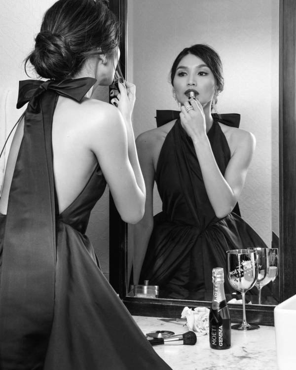 Woman in black halter dress applies lipstick in mirror with champagne and makeup brushes on vanity in black and white portrait.