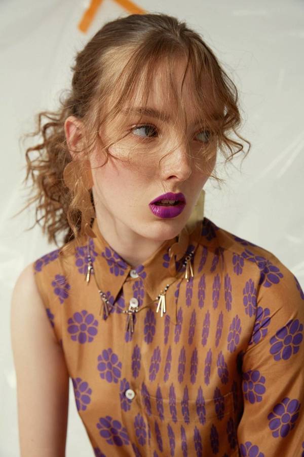 Portrait of a young adult woman with fair skin and curly red hair in a brown and purple patterned shirt, wearing a beaded necklace.