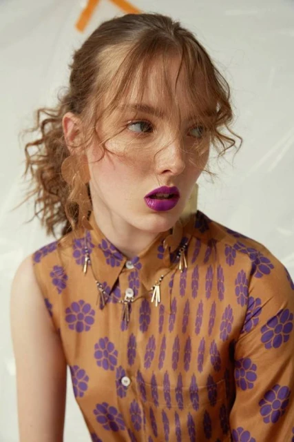 Portrait of a young adult woman with fair skin and curly red hair in a brown and purple patterned shirt, wearing a beaded necklace.