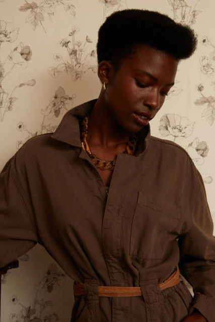 Woman in a brown jumpsuit posing indoors against a floral wallpaper background. She wears a chain necklace and hoop earrings.