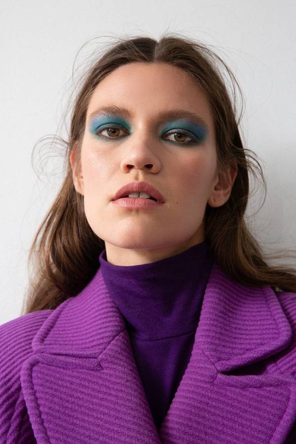Close-up of a fair-skinned adult woman with blue eye shadow and brown hair, wearing a purple turtleneck and coat against a white backdrop.