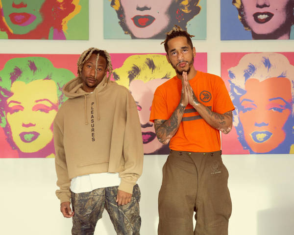 Two men pose indoors against pop art portraits of Marilyn Monroe in front of a white wall, one wearing a tan hoodie and the other an orange t-shirt.