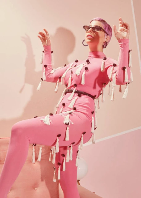 Woman in a pink jumpsuit with white tassels strikes a pose in a photo studio against a pink background. She is wearing black glasses.
