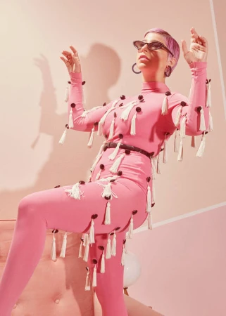 Woman in a pink jumpsuit with white tassels strikes a pose in a photo studio against a pink background. She is wearing black glasses.