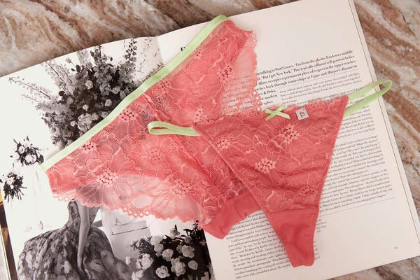 Close-up of two pink lace thong panties with green trim on an open book with black and white floral designs on a tan marble surface.