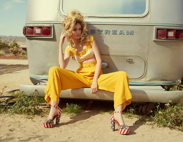 A woman poses on a vintage silver trailer wearing a yellow two-piece outfit and platform heels in an outdoor desert setting.