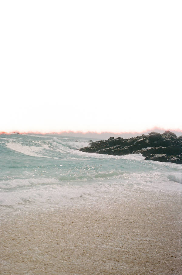 Seascape photograph featuring waves crashing against rocks at the shore with a sandy beach in the foreground against a bright sky.