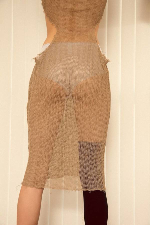 Back view of a slender adult female in a tan-colored knitted midi dress with a slit, posing in front of cream-colored vertical blinds.