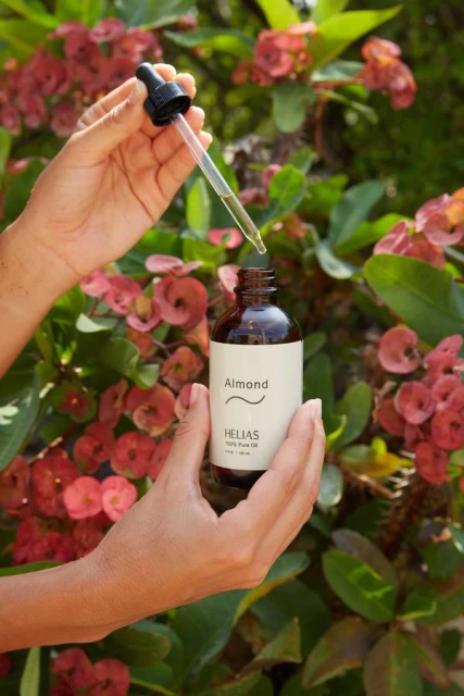 Woman's hands hold a brown glass bottle with a dropper, displaying almond oil in a garden setting with red flowers and green leaves.