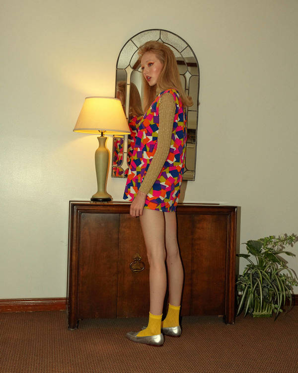 A woman with long red hair stands in a colorful vintage dress, yellow socks, and silver flats, beside a sideboard with a table lamp.