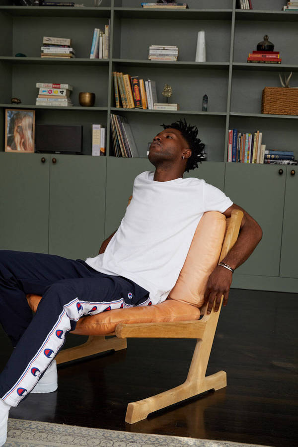 A Black man with dark hair, wearing a white T-shirt and navy track pants, leans back in a wooden chair in an indoor living room setting.