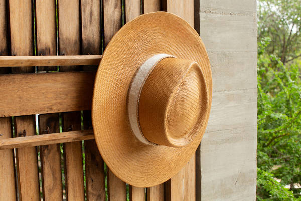 Wide-brim straw hat with light-coloured band hanging against a vertical wooden slat wall and concrete pillar.