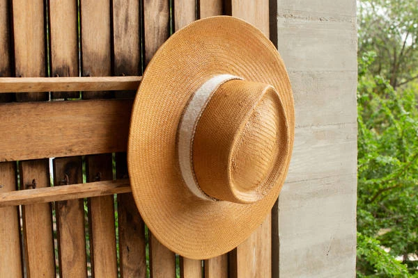 Wide-brim straw hat with light-coloured band hanging against a vertical wooden slat wall and concrete pillar.