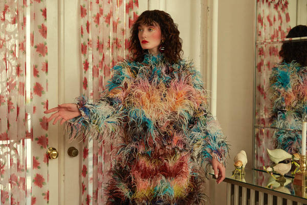 Adult female poses in a vibrant, multi-colored, feathered jacket, indoors, with a mirror reflecting her image and floral wallpaper.