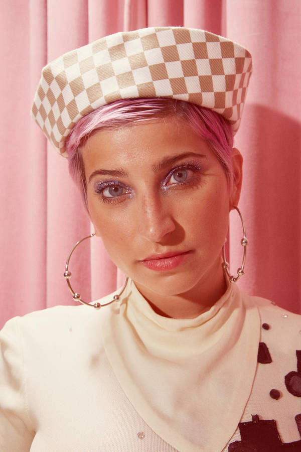 Portrait of a woman with short pink hair wearing a checkered hat and large hoop earrings against a pink curtain backdrop.