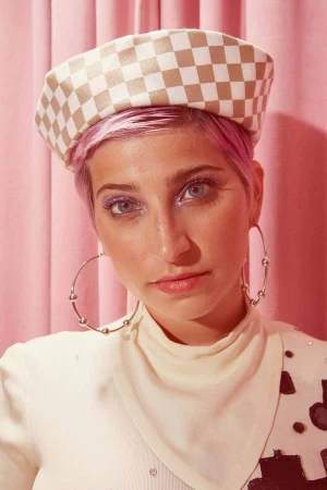 Portrait of a woman with short pink hair wearing a checkered hat and large hoop earrings against a pink curtain backdrop.
